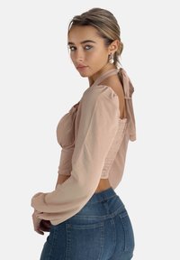 Hellbeige kurz geschnittene Bluse mit langen Ärmeln, gerafftem Rückendesign und einer Schleife am Hals; kombiniert mit dunkelblauen hoch taillierten Jeans.