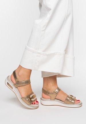 Füße, die goldmetallische Keilsandalen mit Riemen, beigen Sohlen und weißen, gekürzten, weit geschnittenen Hosen tragen.