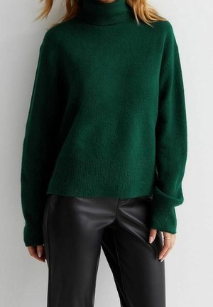 Femme portant un pull à col roulé vert foncé associé à un pantalon en cuir noir sur un fond clair uni.
