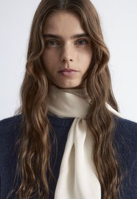 Cremefarbener Seidenschal, der um den Hals gebunden ist, kombiniert mit einem dunkelblauen strukturierten Pullover. Das Haar ist lang, wellig und braun. Einfacher, klarer Hintergrund.