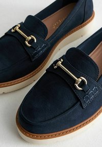 Mocassins en daim bleu marine avec des éléments en or et une semelle plate de couleur claire. Présente un piquage décoratif près du bord supérieur et un design classique.