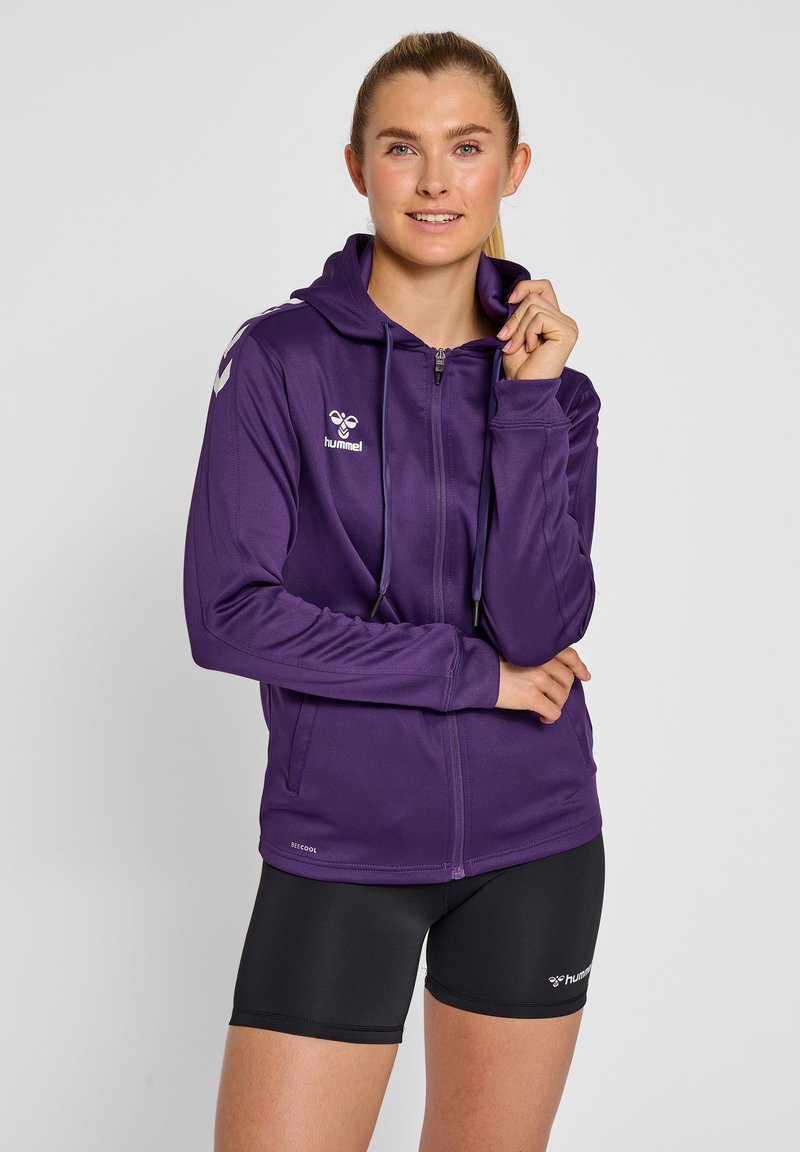 Felpa con zip viola, con cappuccio regolabile, logo sul petto e tessuto leggero. Indossata sopra pantaloni sportivi neri. Tessuto morbido, design atletico.