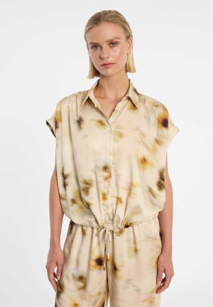 Blonde Frau trägt ein beiges Batik-Top ohne Ärmel mit Knöpfen und einem Tunnelzug in der Taille sowie passende lockere Hosen.