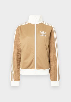 Brązowa kurtka zapinana na zamek błyskawiczny z białymi akcentami i trzema białymi paskami na rękawach. Posiada teksturowany wpleciony wzór na jednym z rękawów. Logo Adidas na klatce piersiowej.