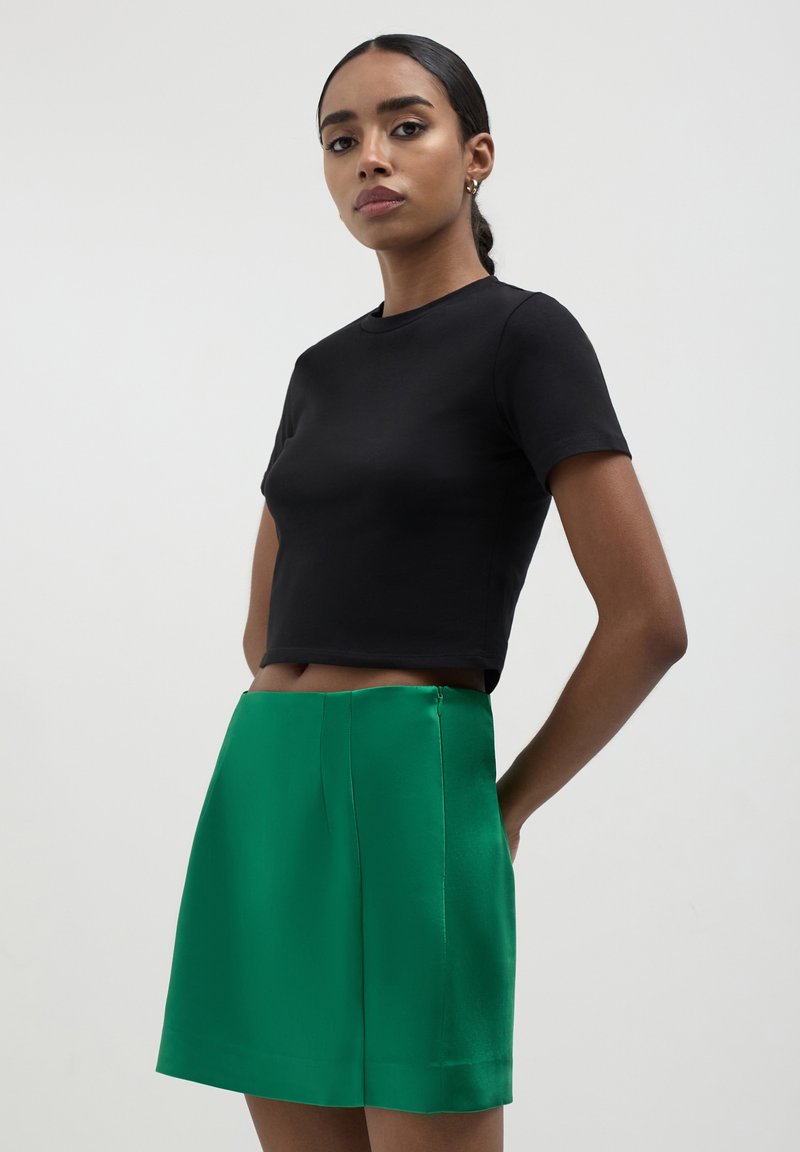 T-shirt noir court à manches courtes associé à une mini-jupe en soie verte présentant une texture lisse et un discret détail de fermeture éclair sur le côté.