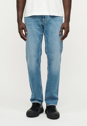 Jasnoniebieskie dżinsy denimowe o prostym kroju nogawki, z efektami wyblaknięcia i tradycyjnym fasonem z pięcioma kieszeniami. Połączone z czarnymi sneakersami na sznurowadła.