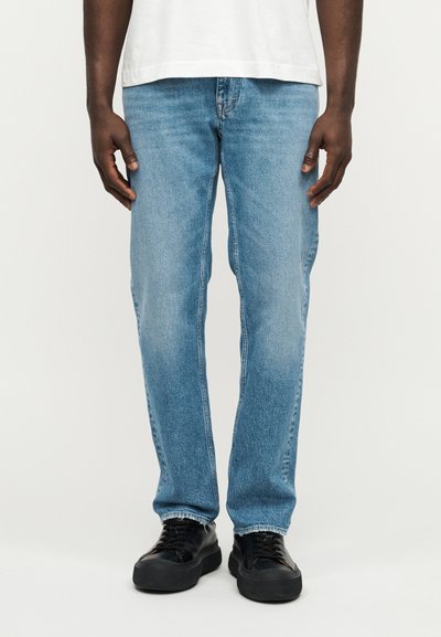 Jean en denim bleu clair avec une coupe droite, des détails délavés et un style traditionnel à cinq poches. Associé à des baskets noires à lacets.