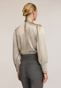Blusa beige setosa con un colletto alto, maniche a sbuffo e un'apertura a occhio di chiave sul retro; abbinata a pantaloni grigi su misura con una vita tessuta.