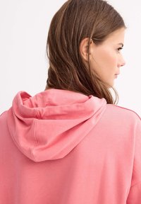 Sudadera rosa con una textura suave y una capucha redondeada, que presenta un corte holgado y detalles de costura visibles en el cuello y las mangas.