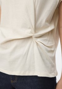 Camiseta de algodón beige con un detalle de nudo retorcido en la espalda, mangas cortas y un ajuste holgado, combinada con jeans de mezclilla azul oscuro.