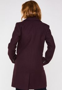 Cappotto in lana bordeaux con una vestibilità su misura, colletto a revers e tasche a patta. Presenta polsini abbottonati e uno spacco posteriore per una maggiore libertà di movimento.