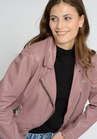 Veste de moto en cuir rose avec des poches zippées et quincaillerie en argent, présentant un design ajusté et une finition texturée. Portée sur un haut noir.