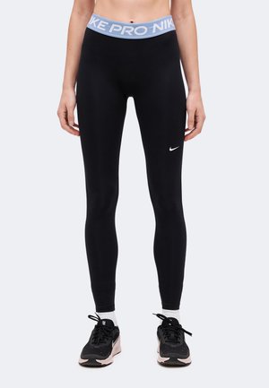 Nő fekete Nike Pro leggingset visel világoskék derékpánttal és fekete Nike futócipővel, fehér zoknival, egyenesen áll.