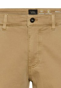 Beige Hosen mit Knopf- und Reißverschluss, ausgestattet mit einem Etikett für "Camel Active" und Icons, die hohe Dehnbarkeit und eine Sicherheitstasche anzeigen.