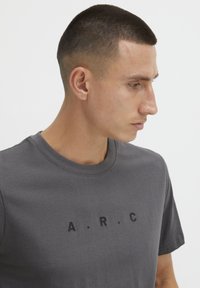 Grå t-skjorte med korte ermer, rund hals og svart "A.R.C."-logo trykket på forsiden, laget av mykt bomullsmateriale.