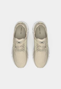 Bézs sportcipők hálós felsőrésszel és fűzőkkel, átlátszó talppal és texturált részletekkel az orr-részen. Belső oldalon látható dombornyomott logó.