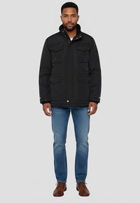 Schwarze gesteppte Jacke mit hohem Kragen, zwei Brusttaschen und Knopfverschluss. Kombiniert mit hellblauen Jeans und braunen Schuhen.