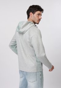 Άνδρας με ανοιχτό γκρι φούτερ με λεπτομέρειες σε μέντα και ανοιχτό μπλε τζιν, γυρισμένος ελαφρώς προς τα πίσω κοιτώντας πάνω από τον ώμο του.