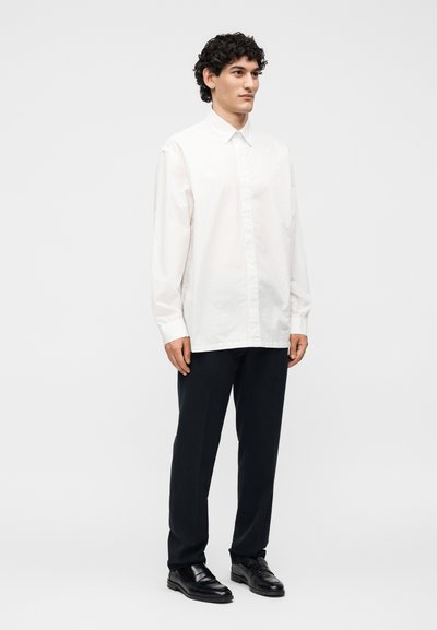 Άνδρας με κατσαρά μαλλιά που φοράει λευκό πουκάμισο με κουμπιά, μαύρο παντελόνι και μαύρα επίσημα παπούτσια, στέκεται μπροστά σε απλό φωτεινό φόντο.