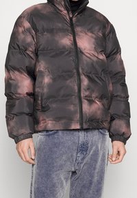 Hombre vestido con una chaqueta acolchada de teñido anudado en negro y rosa, con cremallera frontal y bolsillos laterales, combinada con pantalones de pana azul claro.