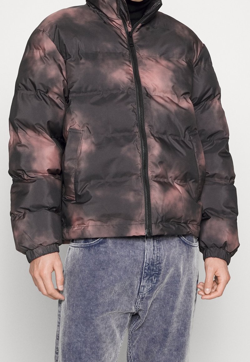 Hombre vestido con una chaqueta acolchada de teñido anudado en negro y rosa, con cremallera frontal y bolsillos laterales, combinada con pantalones de pana azul claro.