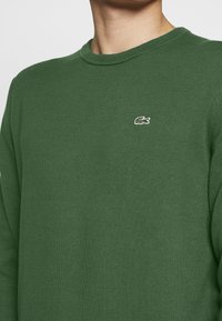 Pullover verde in maglia con colletto a girocollo e un piccolo logo ricamato sul petto. La texture è liscia e la forma è aderente.