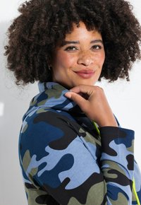 Giacca con cappuccio in un motivo camouflage blu, verde e nero. Realizzata in tessuto morbido, presenta una vestibilità comoda e un laccio contrastante.