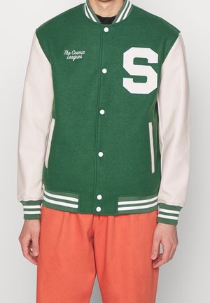 Mężczyzna w zielono-kremowej kurtce varsity z literą „S” i napisem „The Cosmic Leagues”, w pomarańczowych spodniach, stojący na białym tle.