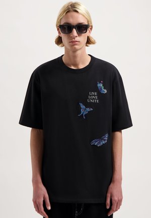 Zwarte t-shirt met korte mouwen, voorzien van geborduurde kleurrijke vlinderontwerpen en de tekst "LIVE LOVE UNITE" op de voorkant.