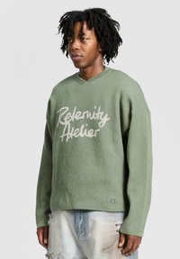 Groene gebreide sweater met een losse pasvorm, voorzien van een v-hals en witte geborduurde tekst "Reelenity Atelier" op de voorkant, met een gestructureerde stof.