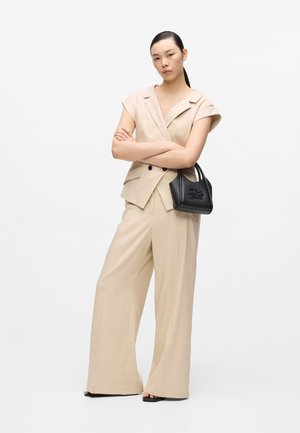 Žena u bež prsluku bez rukava i širokim hlačama, drži mali crni ručni torbu, stoji s prekriženim rukama na bijeloj pozadini.