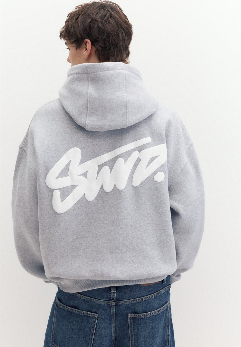 Jeune personne aux cheveux courts et bruns, portant un sweat à capuche gris avec un texte blanc stylisé dans le dos et un jean bleu, vue de dos.