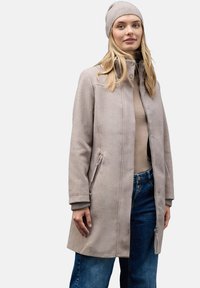 Cappotto in misto lana color beige chiaro con colletto alto, zip frontale, tasche laterali e polsini a coste. Abbinato a un berretto di maglia coordinato.