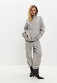 Lichtgrijze hoodie met een ritssluiting aan de voorkant en grote zakken, gecombineerd met bijpassende grijze joggers en zwarte puntige laarzen, wat een gezellige uitstraling creëert.
