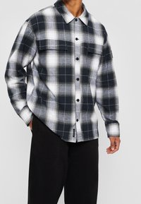 Camicia in flanella a quadri bianco e nero con due tasche sul petto, chiusura con bottoni, maniche lunghe e orlo leggermente curvo. Tessuto strutturato.