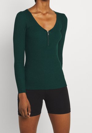 Dark green ribbed long-sleeve top with a deep V-neck and a front zipper, featuring a fitted design and textured fabric. Worn with black shorts.