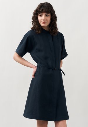 Frau mit lockigem dunklem Haar, die ein kurzärmeliges marineblaues Kleid mit einem dünnen Gürtel trägt, steht mit einer Hand auf der Hüfte vor einem schlichten Hintergrund.