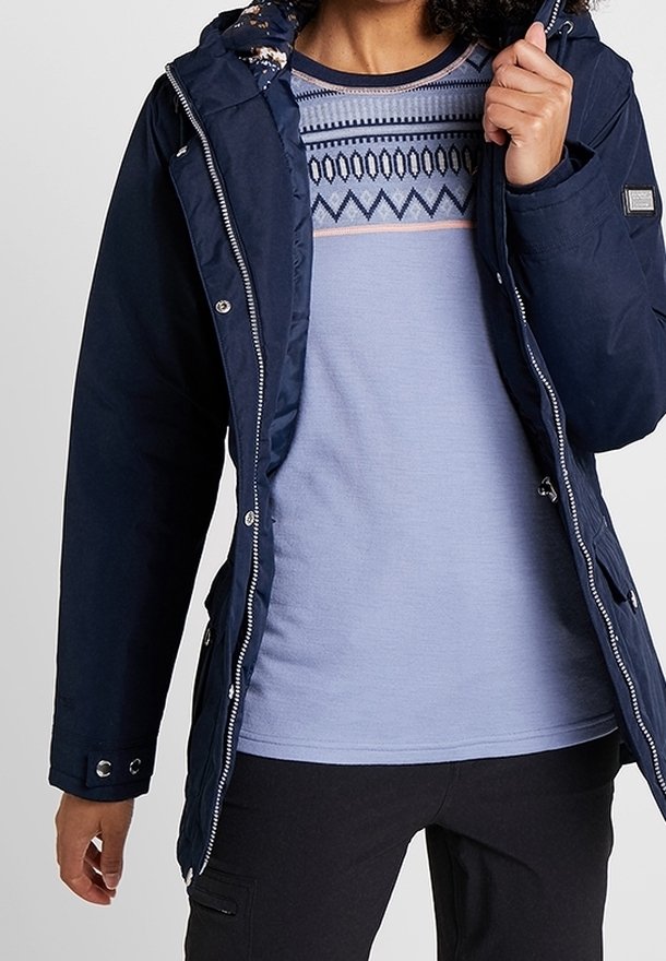 Veste isolante bleu marine avec capuche, fermeture éclair à l'avant, pressions argentées et doublure florale ; superposée sur un haut à motifs bleu clair et un pantalon noir.