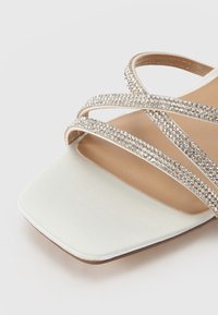 Sandalo piatto crema con cinturini incrociati adornati da strass trasparenti. Texture liscia e design a punta quadrata. Suola piatta di colore beige.