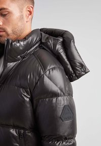 Giubbotto in piuma nero con una finitura lucida, dotato di un alto colletto, tasche laterali con zip e un badge in gomma sul maniche.