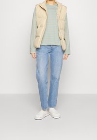 Beige puffig väst över en randig blå långärmad skjorta, tillsammans med ljusblå jeans och vita slidesandaler. Enkel, avslappnad outfit.