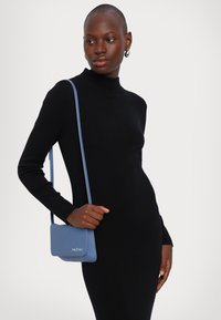 Sac à épaule en cuir bleu doux avec une finition texturée, doté d'une fermeture à rabat et d'un logo embossé. Modèle portant une robe noire ajustée.