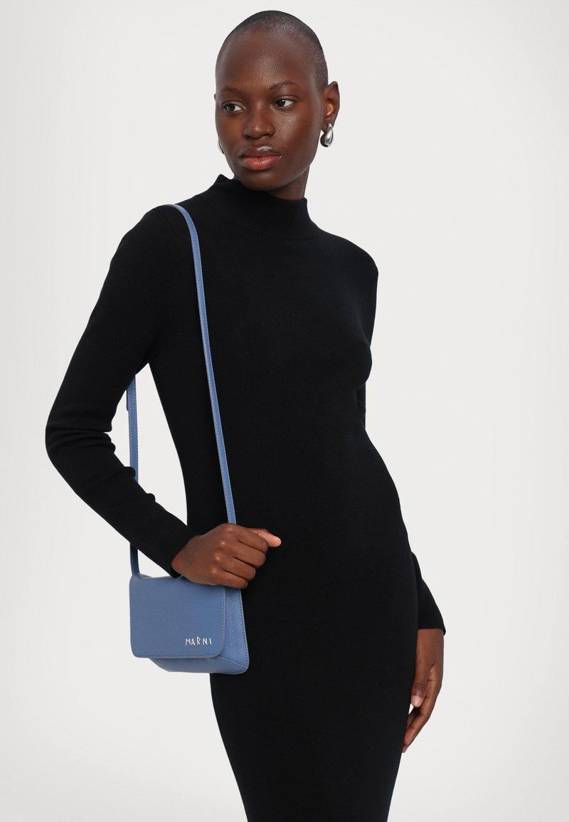 Borsa a spalla in morbida pelle blu con finitura testurizzata, dotata di chiusura a patta e logo impresso. Modella in un abito nero aderente.