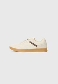Basket en crème avec une tige texturée, une bande d'accent brun et une semelle en caoutchouc beige, présentant un profil bas et un design à lacets.