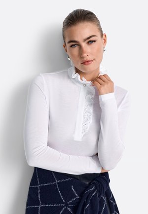 White long-sleeve shirt with a ruffled collar and button detail, paired with a dark blue knit sweater tied at the waist featuring a diamond pattern.