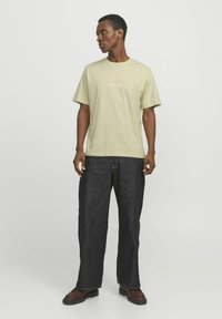 Helle grüne, kurzärmlige T-Shirt mit dem Aufdruck "Originals Studio", kombiniert mit dunkelblauen, weit geschnittenen Jeans und braunen Stiefeln. Einfaches, lässiges Design.