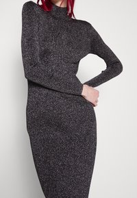Robe noire brillante à côtes avec accents métalliques, col haut, manches longues et coupe ajustée mettant en valeur la texture et la silhouette.