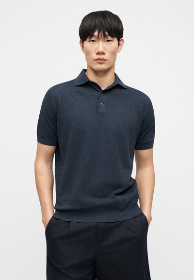Μπλε σκούρο πόλο μπλουζάκι κατασκευασμένο από απαλό πλεκτό ύφασμα, με κλασικό γιακά, κοντά μανίκια και πατιλέτα με τρία κουμπιά. Συνδυάζεται με σκούρο παντελόνι.