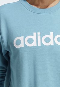 Ljusblå sweatshirt i mjukt tyg, med en stor vit "adidas"-logga. Ribbad krage och muddar. Slät yta med en avslappnad design.