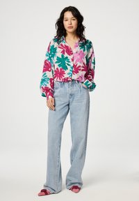 Floral button-up shirt in pink, green, and teal on light fabric, paired with light blue high-waisted denim jeans and pink slip-on shoes.